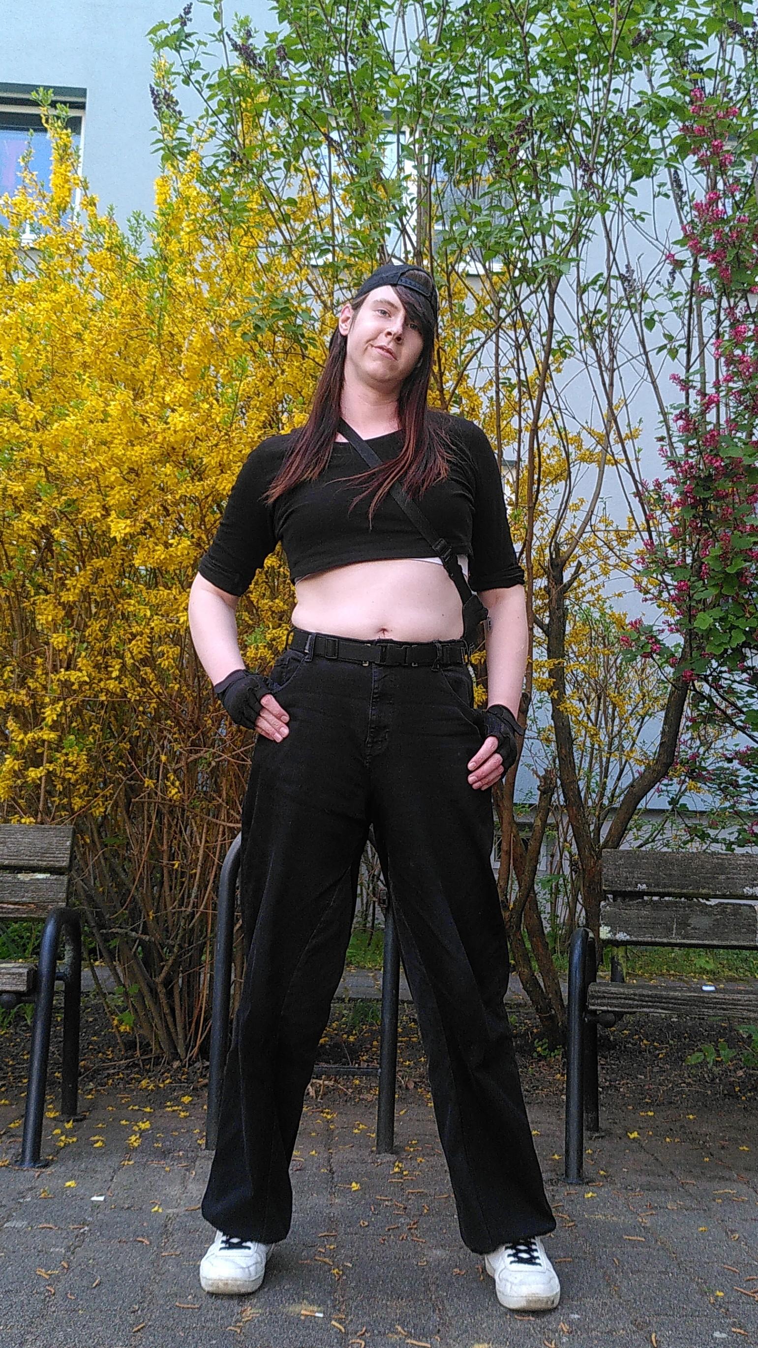 Picture of Sam (🔥) standing in front of some bushes that are blooming in yellow and cerise. 
He's wearing a black, cropped long sleeve, rolled up to his elbows, black, high rise baggy pants, white sneakers with black laces, a black base cap, visor backwards with a "Sabotage" patch and black fingerless gloves. Their white sports bra is peaking out under the crop top. He also has a fanny pack, which is slung over their shoulder. 
They look very self confident.